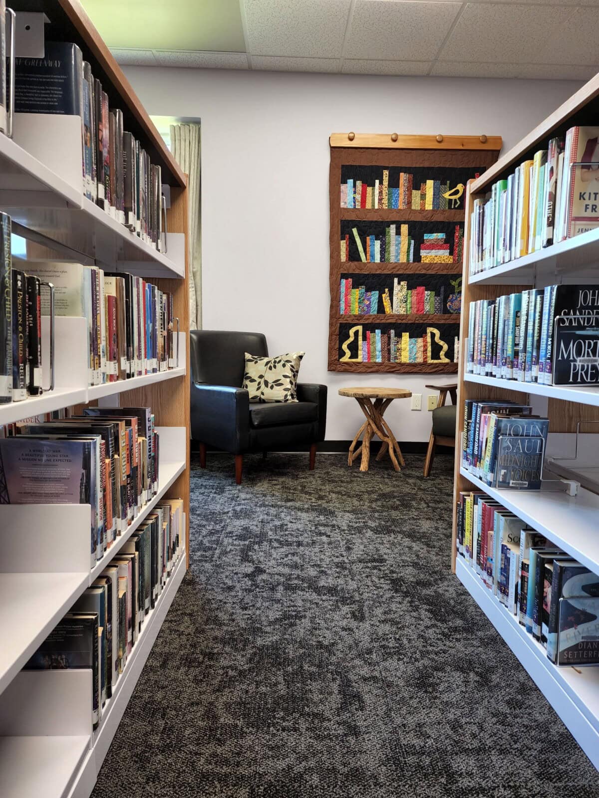 A cozy library corner with two bookshelves, a black armchair with a patterned pillow, a small wooden table, and a decorative quilt on the wall depicting a bookshelf filled with colorful books.
