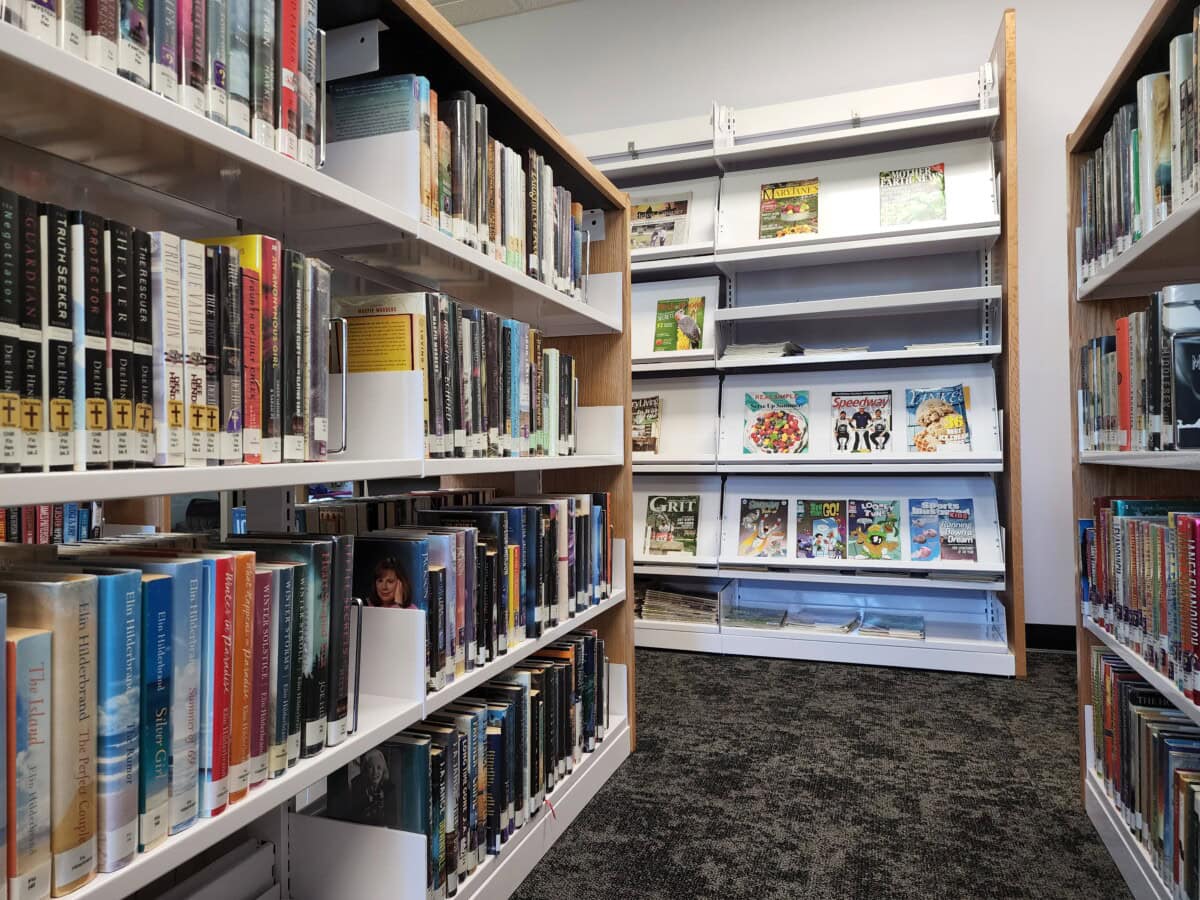 Bookshelves filled with books line both sides of a library aisle, leading to a rack displaying colorful magazines and picture books. The carpeted floor and bright lighting create a welcoming atmosphere.
